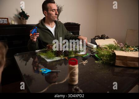 A man decorates festive Christmas decorations using pine cones, pine branches, twine, and glue on a dining table in a cozy home setting, conveying war Stock Photo