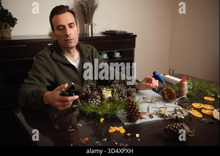 A man sits at a dining table crafting a festive centerpiece with pine cones, greenery, twine, and oranges using a hot glue gun in a cozy home setting. Stock Photo