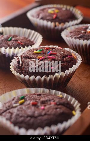close-up of baked chocolate muffins with multi-colored sprinkles, fresh brownies in white paper liners on brown wooden tray, dramatic lighting in soft Stock Photo