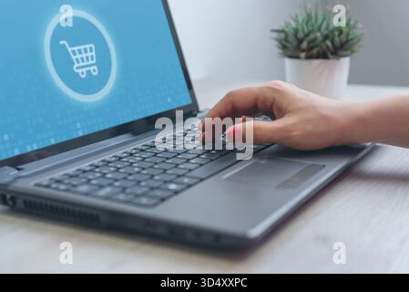 Person typing on laptop with blue shopping cart icon on screen, symbol of online store, e-commerce, and digital purchase process Stock Photo
