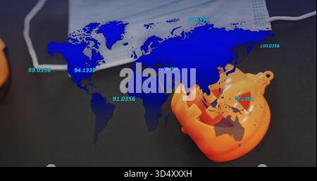 Showing pale blue world map overlaying dark desk, with teal labels, surgical mask, pumpkin ornament Stock Photo