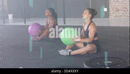 Sitting partners holding green, purple medicine balls on rubber mat, wearing sports bras and shorts Stock Photo