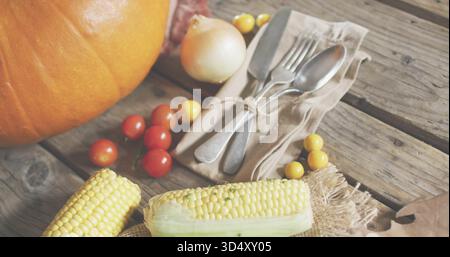 Showing partly husked corn resting on wooden plank, with pumpkin, tomatoes, onion and tied cutlery Stock Photo