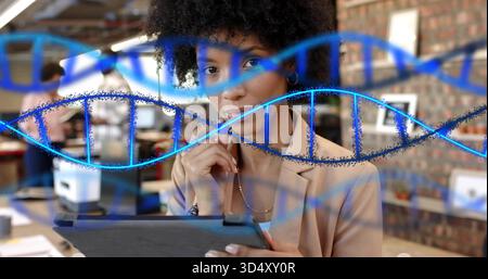 Reading woman in beige blazer touching chin while holding tablet in open office, with DNA overlay Stock Photo