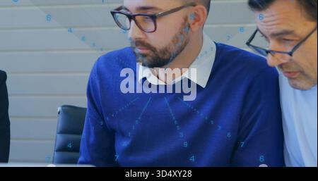 Reviewing bearded man in blue sweater and glasses leaning over laptop at office, with documents Stock Photo