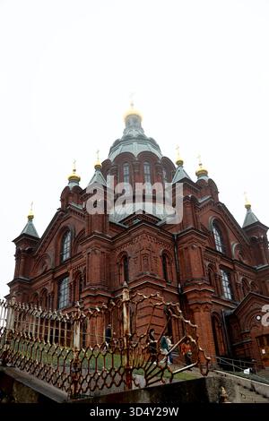 Brick facade and domes of Uspenski Cathedral in Helsinki, Finland, viewed past an ornate iron fence with visitors on the steps under a foggy sky. Stock Photo