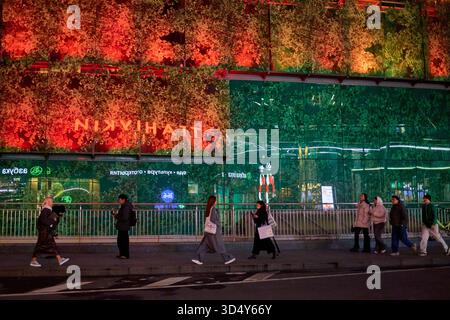 People walking down the street in front of colorfully lit glass paneled wall at night. Moscow City, Moscow, Russia. Stock Photo