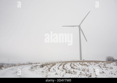 View of a solitary wind turbine stands tall against a muted sky, contrasting with the snow-covered field in winter, Syracuse, New York, United States. Stock Photo