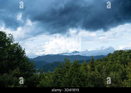 View of a mountain range shrouded in mist under a dramatic sky, framed by lush green trees, creating a serene yet powerful landscape, Takayama, Gifu, Stock Photo