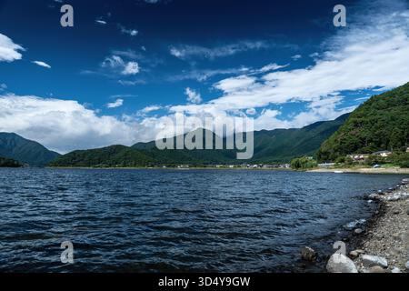 View of the majestic, dark mountain range reflected on the rippling lake under a vibrant, blue sky dotted with fluffy white clouds, Fujikawaguchiko, Y Stock Photo