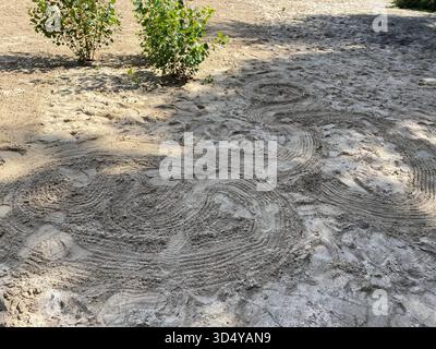 Artistic swirl patterns drawn in the sand of a sunny park or garden area, with small green bushes nearby. Stock Photo