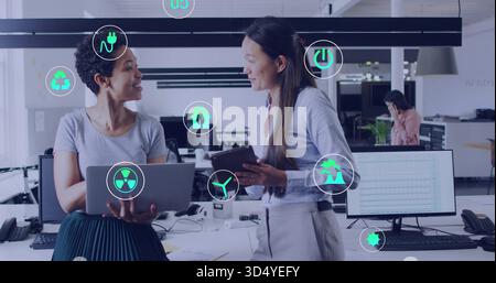 Talking two women in workwear holding laptop tablet in open office showing energy icons Stock Photo
