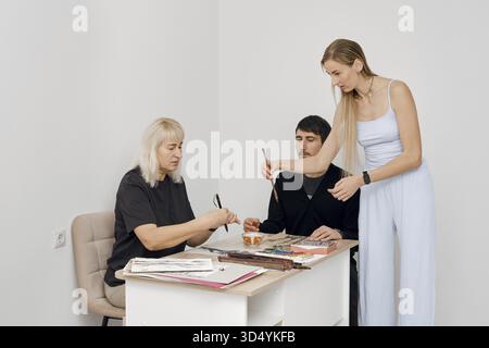 Three individuals engage in a creative workshop, sharing art supplies and discussing their work. The bright room provides a welcoming space for creati Stock Photo