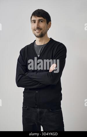 A man is posing confidently with arms crossed, wearing a black cardigan over a gray shirt. He stands against a simple, light backdrop, giving a relaxe Stock Photo