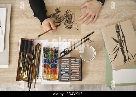 A person is painting with watercolors and sketching with fine brushes on brown paper. Art supplies are organized on a wooden table, including paints, Stock Photo