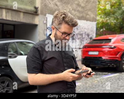 Man using smartphone on city street near parked cars. Technology, urban lifestyle, and everyday communication in modern environment. Stock Photo