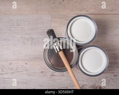 Three cans of paint and a wooden-handled paintbrush lie on a light wood grain surface. Stock Photo