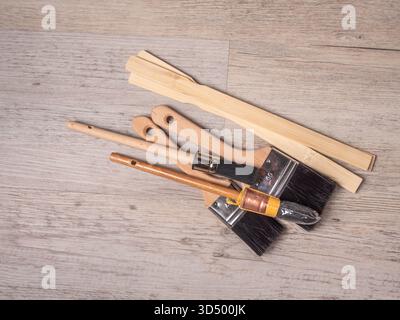 A pile of assorted paintbrushes and wooden stir sticks rests on a light gray-brown wood grain surface. Stock Photo