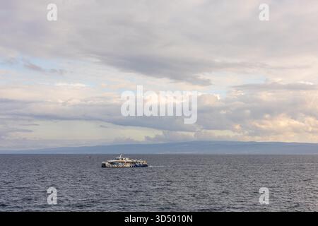 A lone catamaran glides across a vast sea under a soft, overcast sky, with distant land on the horizon. The scene conveys calm, travel, and maritime s Stock Photo