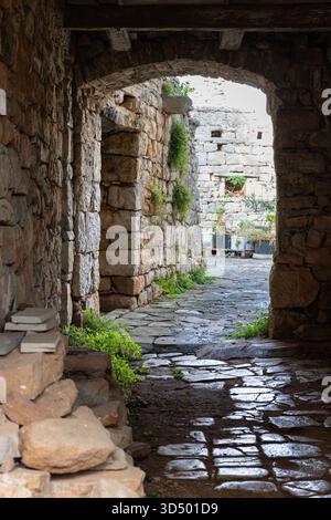 A narrow stone arch opens onto a sunlit courtyard in an old village. Weathered walls, greenery, and a winding cobblestone path create a rustic, tranqu Stock Photo