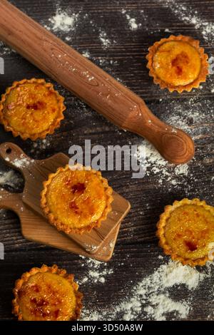 Tarts fresh from the oven sit on a wooden board. A rolling pin and flour dust the dark kitchen table, capturing a moment of baking joy. The warm color Stock Photo