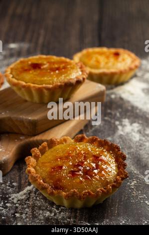 Mini lemon tarts with a golden top are placed on a wooden board. The surface is dusted with flour, creating a cozy kitchen vibe that highlights the ha Stock Photo