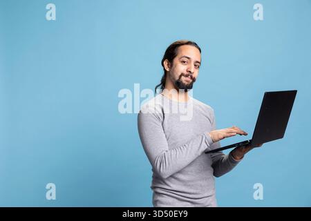 Confident thoughtful man working on his laptop while engaging in digital communication, standing against blue background. Young casual adult doing remote work on modern online platforms. Stock Photo
