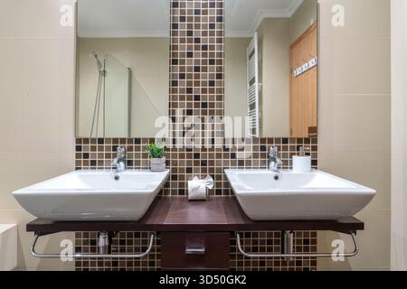 Stylish bathroom showcases two white sinks on a wooden counter, complemented by square mosaic tiles and double mirrors, creating a contemporary look Stock Photo