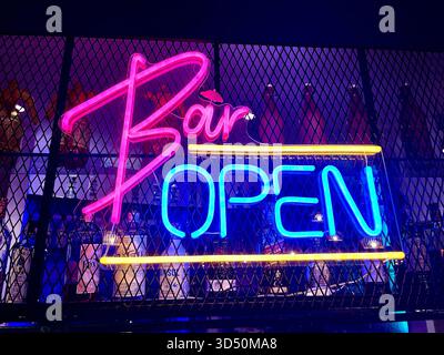 Neon “Bar OPEN” sign glows in pink, blue, and yellow against mesh and bottles—inviting guests into a vibrant nightlife scene Stock Photo