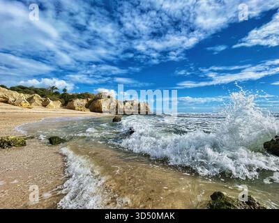 Foamy waves crash on the rocks of the secluded Praia dos Bés, Albufeira, Algarve, Portugal, beneath cliffs and a vivid sky scattered with clouds Stock Photo
