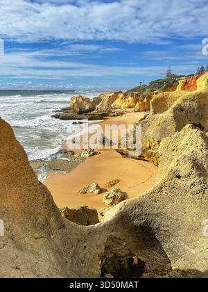 Foamy waves crash on the rocks of the secluded Praia dos Bés, Albufeira, beneath cliffs and a vivid sky scattered with clouds Stock Photo