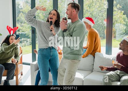 Diverse friends singing karaoke in home living room, wearing festive hats, holding microphones Stock Photo