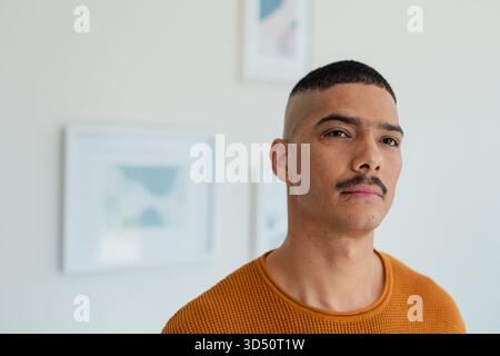 Man wearing orange sweater standing against pale wall at home with abstract art prints, copy space Stock Photo