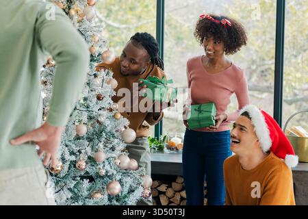 Diverse friends gathering around flocked Christmas tree in living room, holding green gift boxes Stock Photo