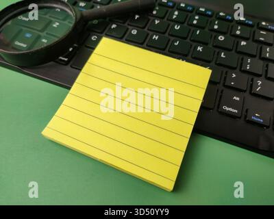 The image displays a pad of yellow, lined sticky notes resting on a black computer keyboard, next to a magnifying glass. Stock Photo