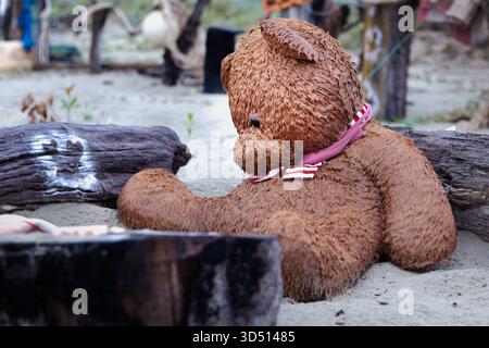 A large, saturated brown teddy bear with a pink and white striped scarf sits despondently or contemplatively in the wet sand on a beach Stock Photo
