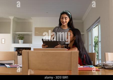 Collaborating diverse female friends using laptop and tablet at home dining area, with wooden table Stock Photo