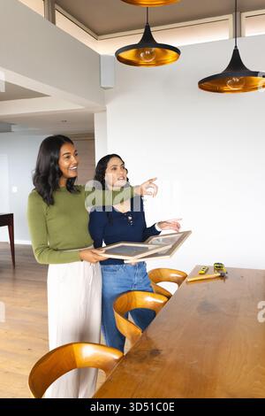 Diverse female friends planning picture frame placement in modern dining room, with tape measure Stock Photo