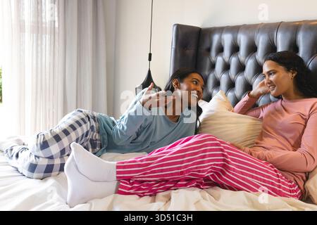 Reclining diverse female friends conversing on bed in cozy bedroom, with pillows and pendant lamp Stock Photo