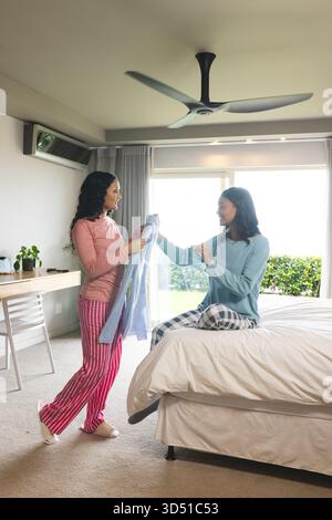 Diverse female friends passing pastel blue cardigan in modern bedroom, with smartphone on desk Stock Photo
