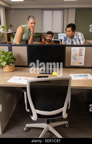 Leaning Diverse coworkers examining printed charts at cubicle desk, with large flat-screen monitor Stock Photo