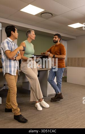 Talking Diverse coworkers standing near workstation in open-plan office, with coffee cup and tablet Stock Photo