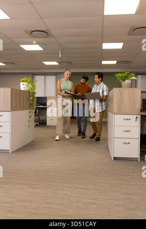 Walking Diverse coworkers discussing binder and laptop in office aisle, with notebook and plants Stock Photo