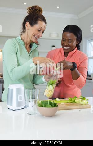 Diverse female friends mixing green smoothie in home kitchen, with blender jar and spinach bowl Stock Photo