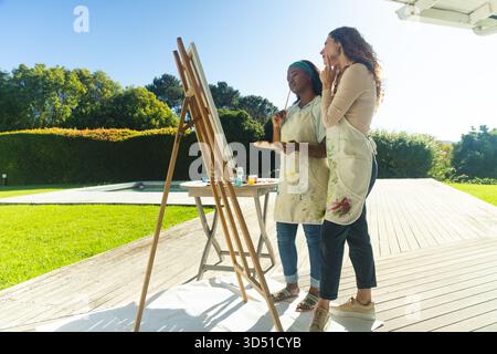 Holding brushes Diverse female friends examining canvas on easel on wooden deck, with paint jars Stock Photo
