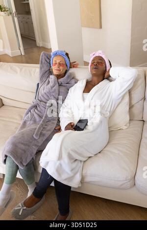 Relaxing Diverse female friends reclining on cream-colored sofa in living room, with smartphone Stock Photo
