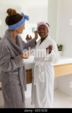 Diverse female friends applying makeup in home bathroom, with bathrobes and vanity mirror Stock Photo
