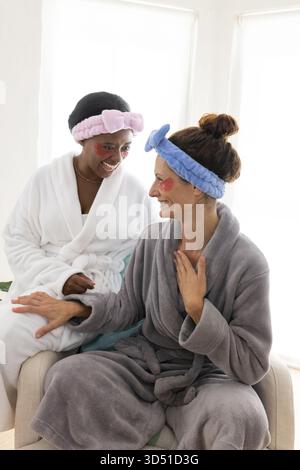 Chatting Diverse senior women friends sitting on sofa in living room, with bathrobes and headbands Stock Photo