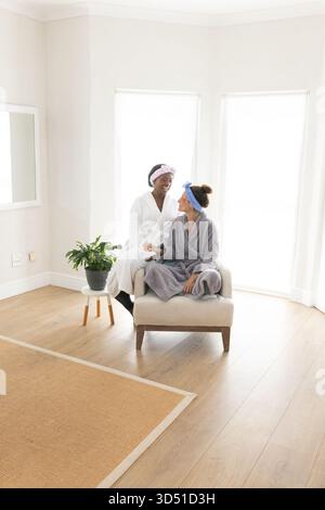 Smiling Diverse female friends sitting on bench in minimalist room, with bathrobes and plant Stock Photo