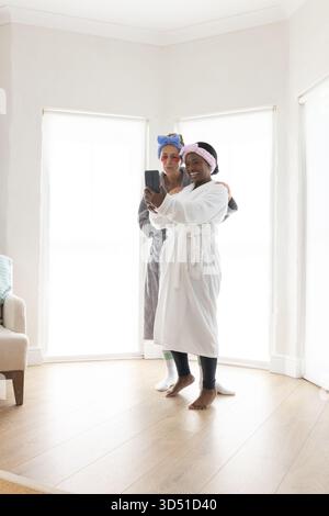 Taking selfie Diverse female friends wearing plush bathrobes in spa corner of home, with smartphone Stock Photo